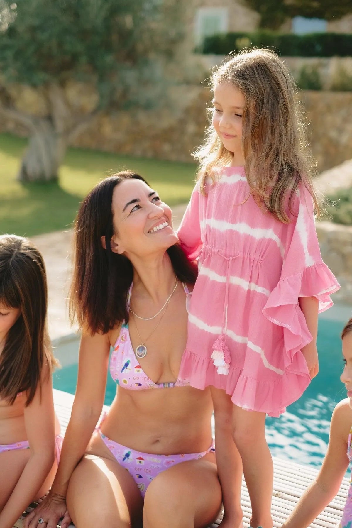 Mother in pink bikini smiling at daughter wearing pink tie-dye caftan by the poolside