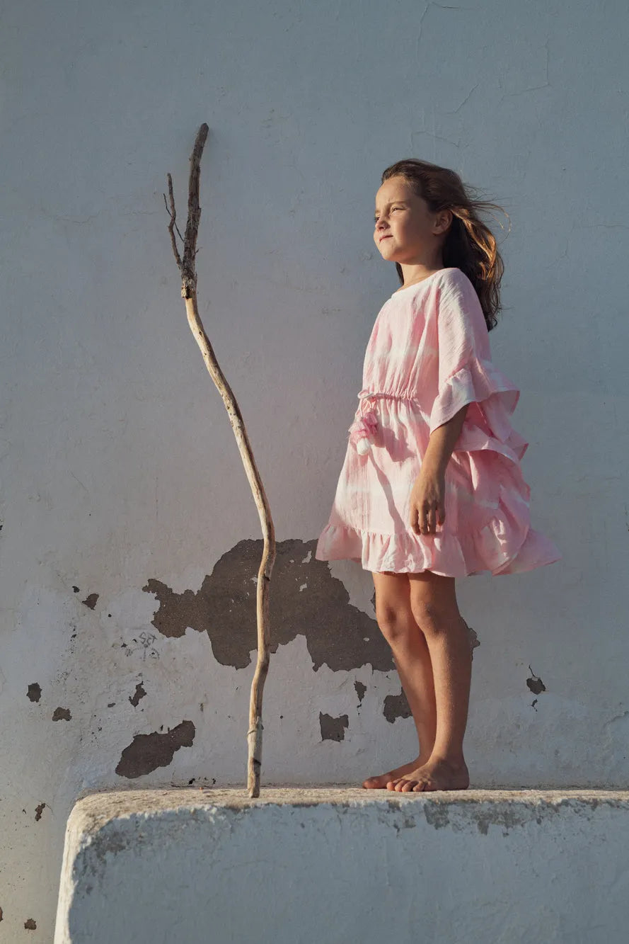 Young girl standing barefoot in pink ruffled dress near weathered wall and tall stick outdoors