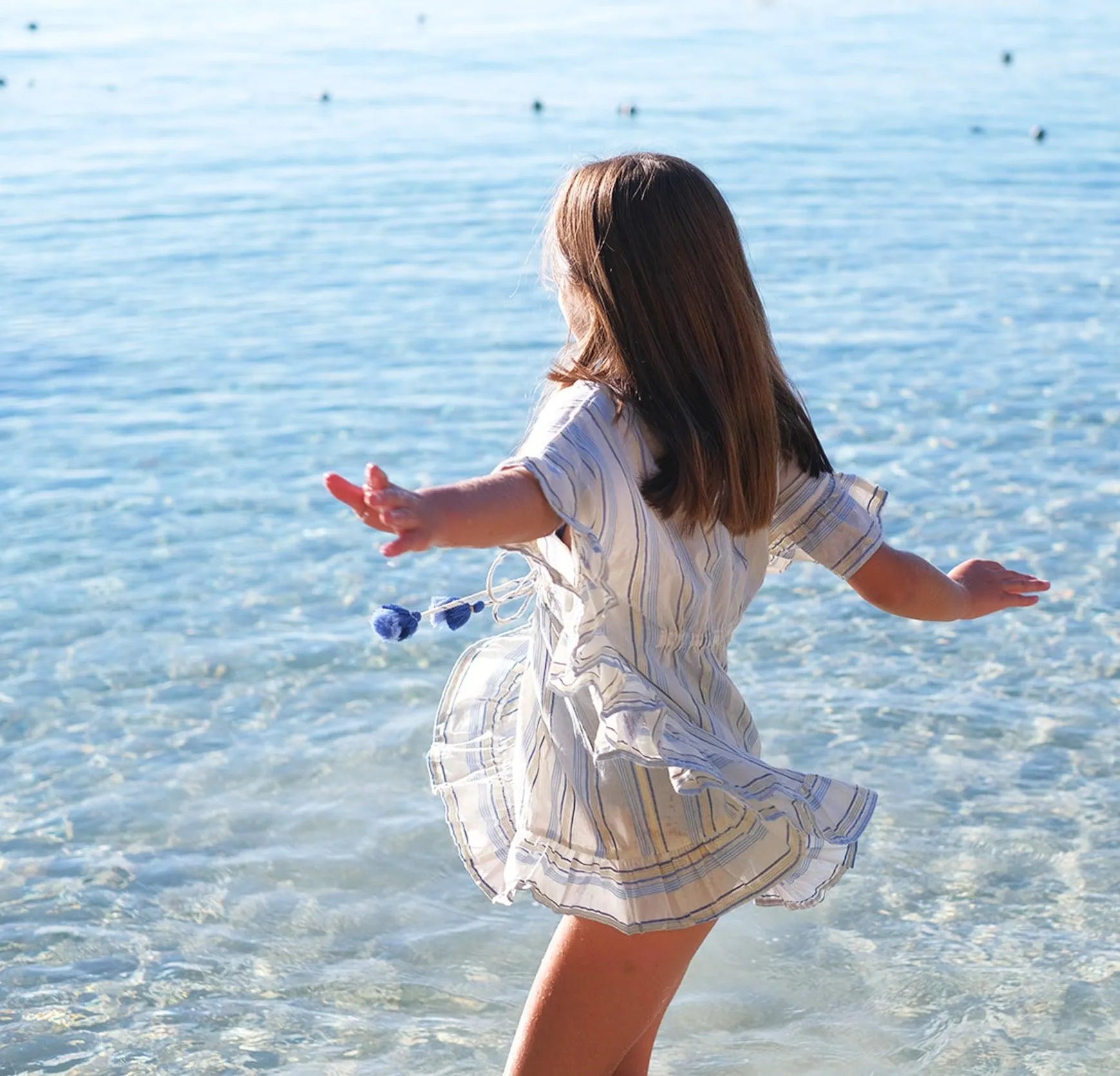 Girl spinning by clear ocean water wearing white and blue striped dress with tassels