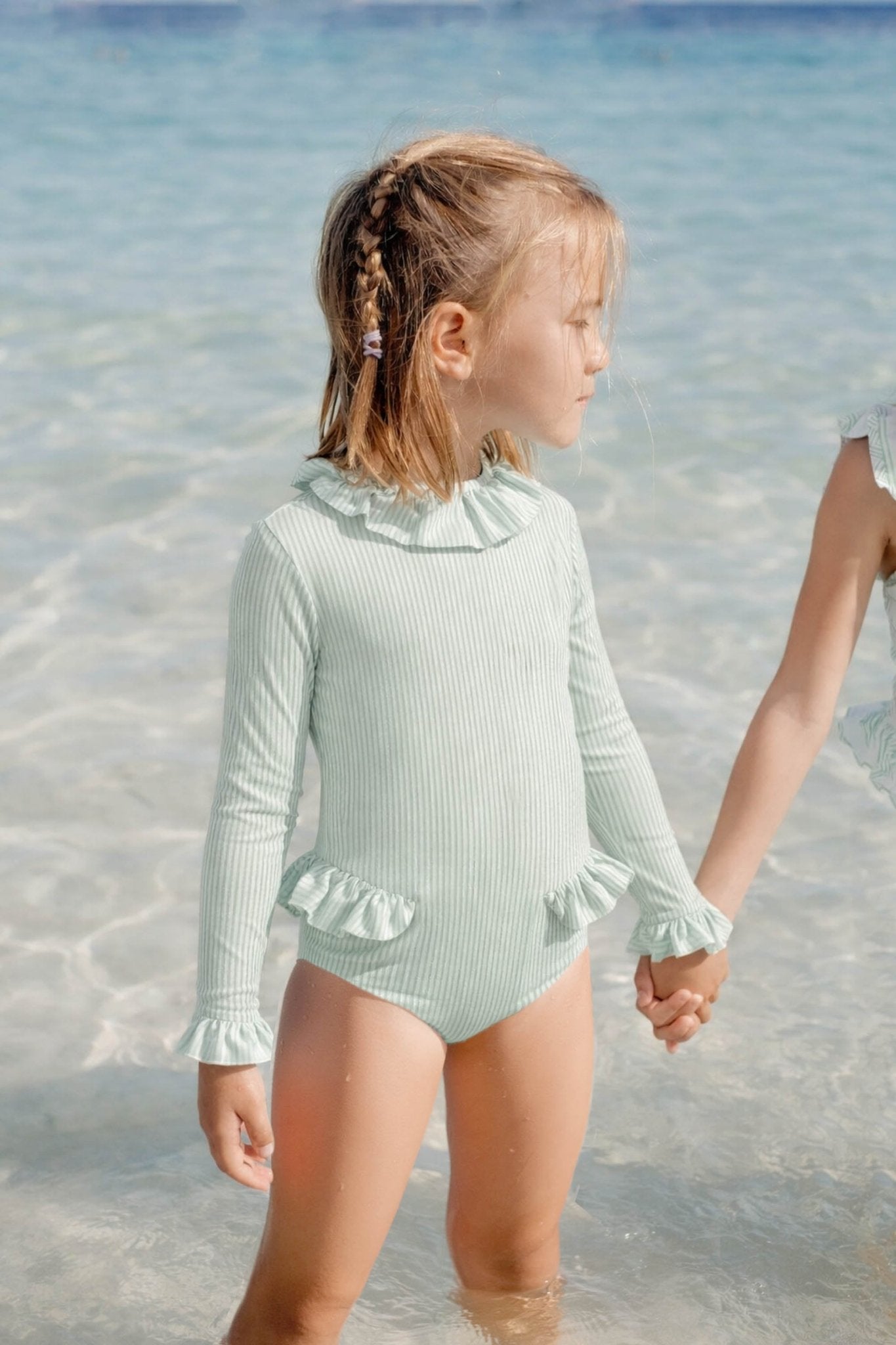 Girl in pastel green long-sleeve ruffled swimsuit standing in shallow beach water