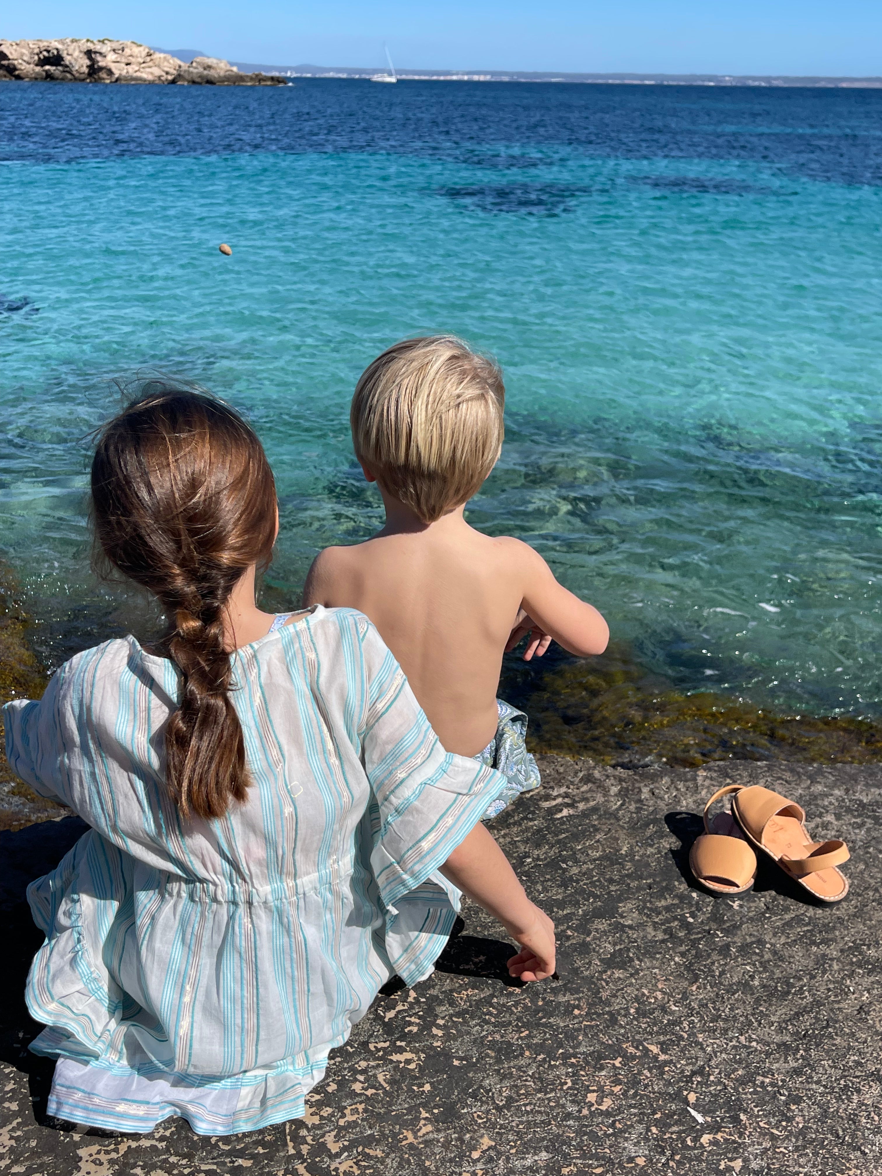 Two children sitting on rocky shore by turquoise sea, one wearing striped dress, sandals nearby