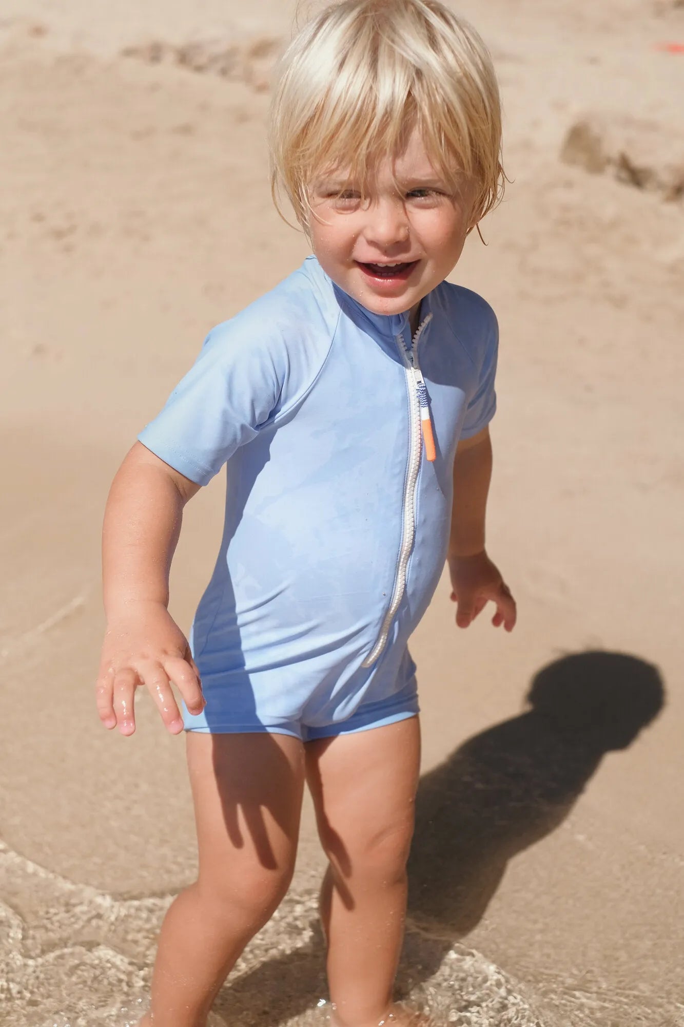 Toddler boy in light blue short-sleeve UV protection swimsuit playing at sandy beach