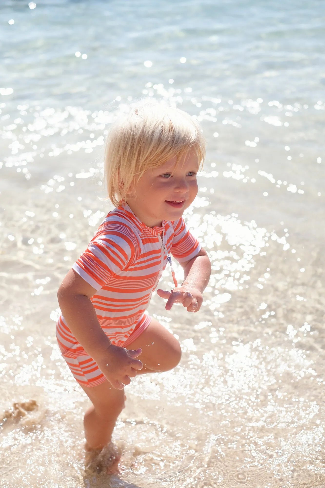 Toddler boy playing in shallow water wearing short-sleeve orange striped UV protective swimsuit