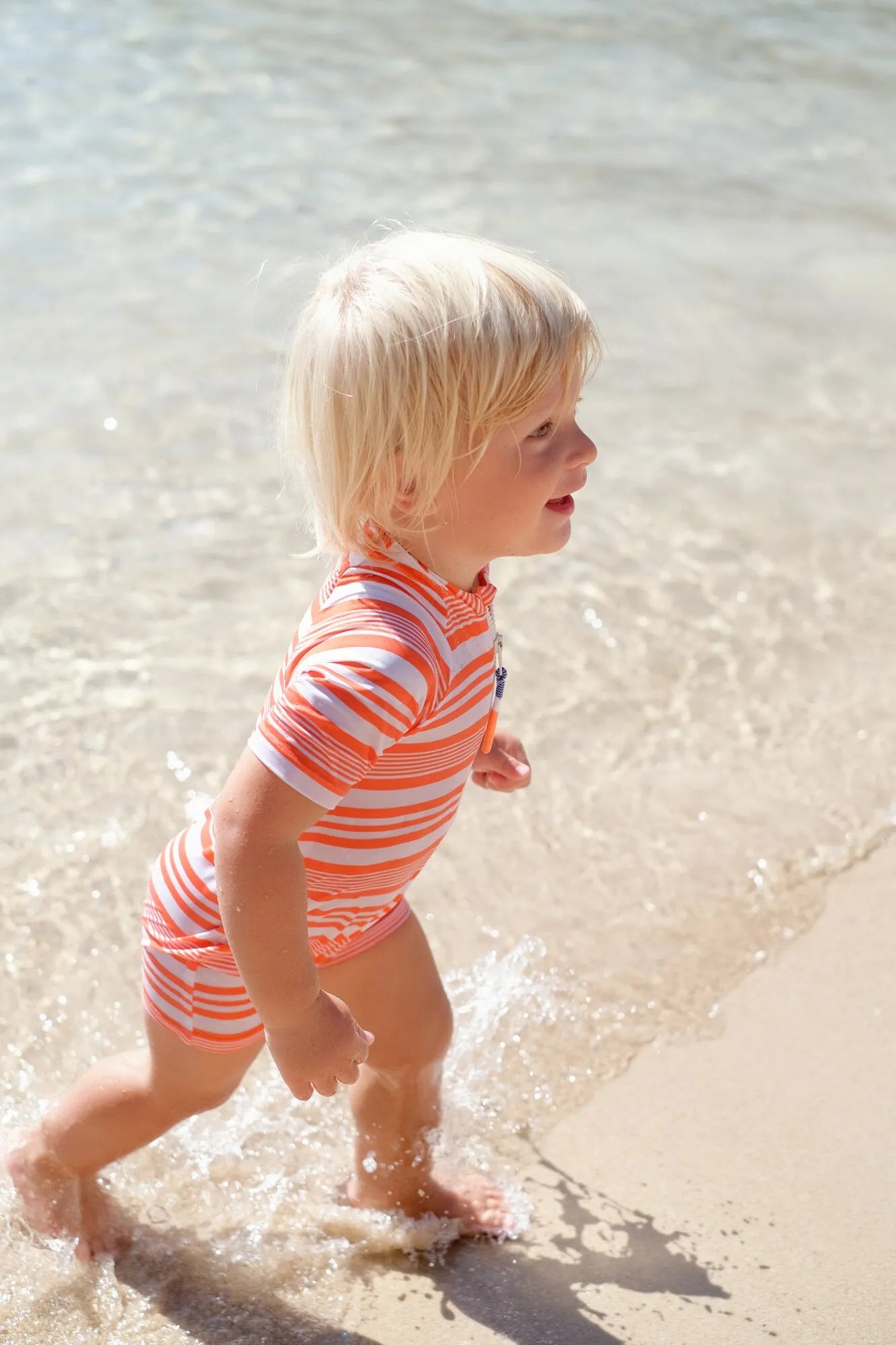 Toddler boy wearing orange striped short-sleeve UV protective swimsuit playing at beach shore
