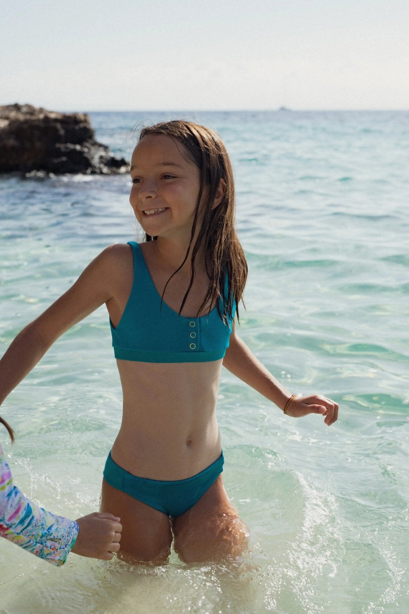 Girl wearing teal two-piece UV swimwear smiling in the sea, with rocky shore in background