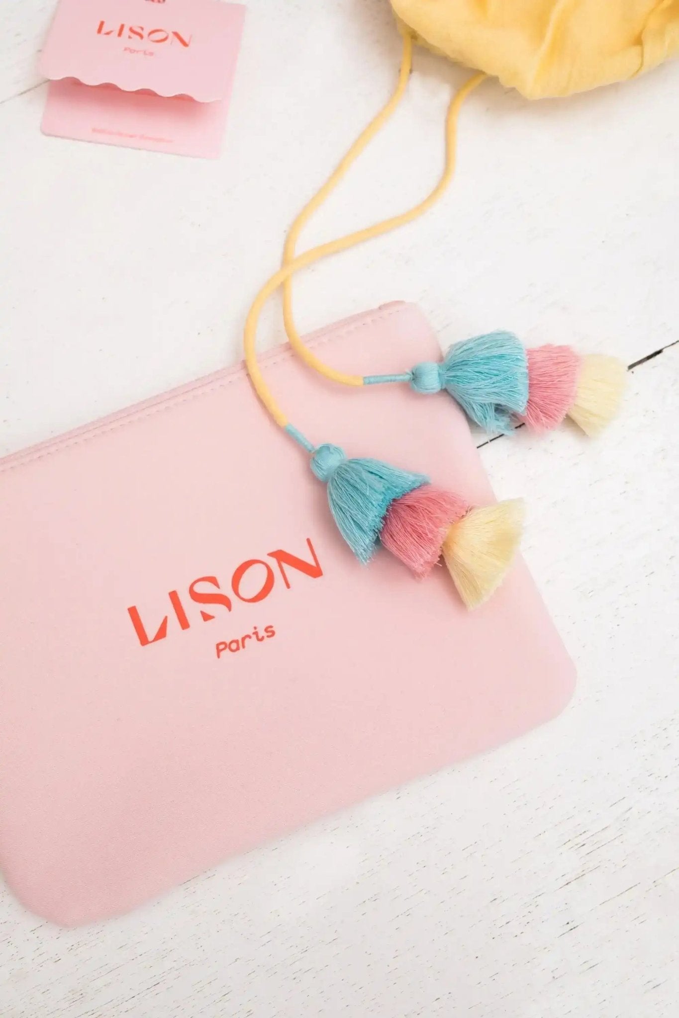 Pastel pink pouch with 'Lison Paris' logo, yellow cord, and colorful tassels on white wood