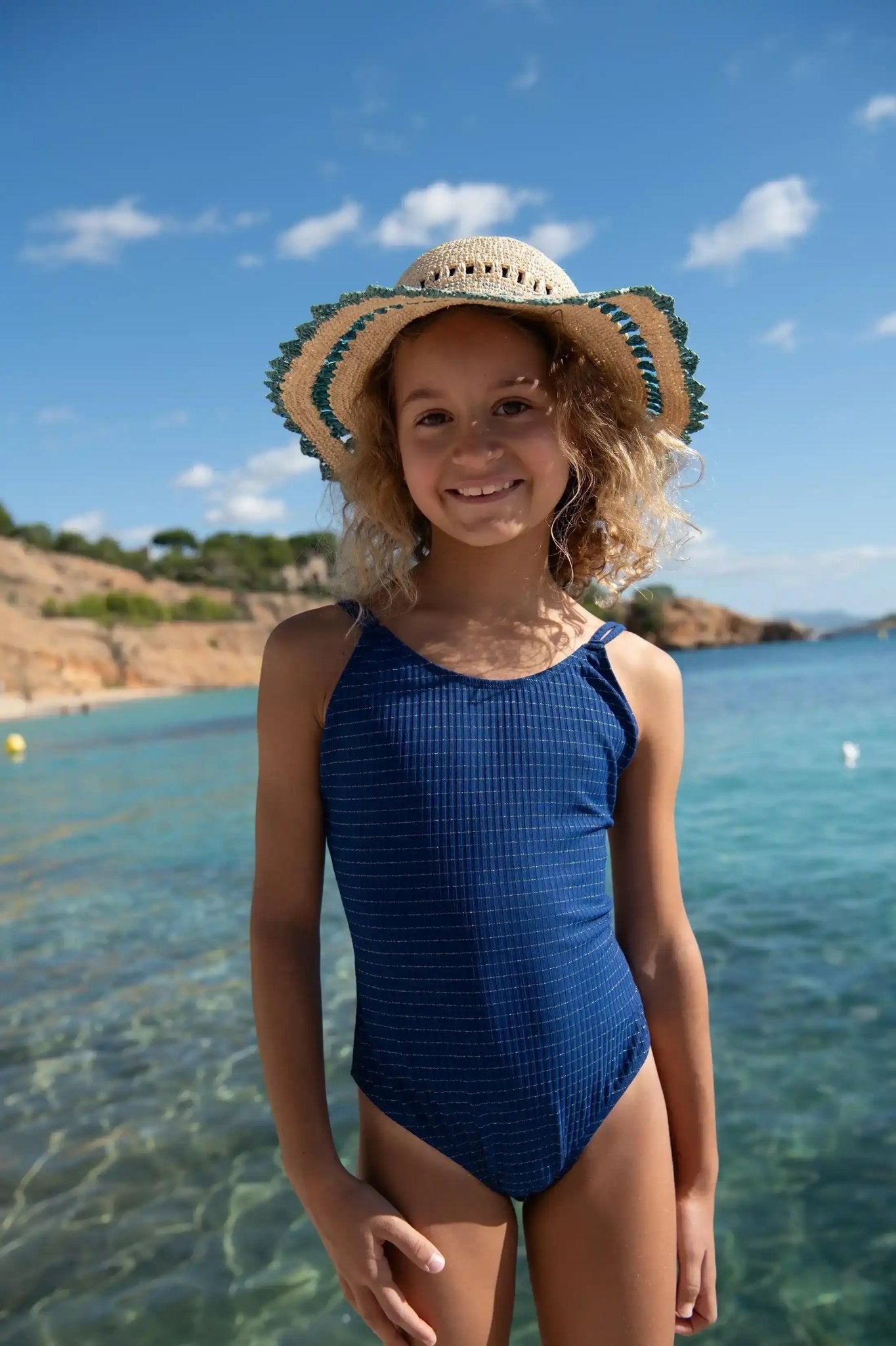 Girl in navy blue one-piece swimsuit and straw hat smiling at the beach