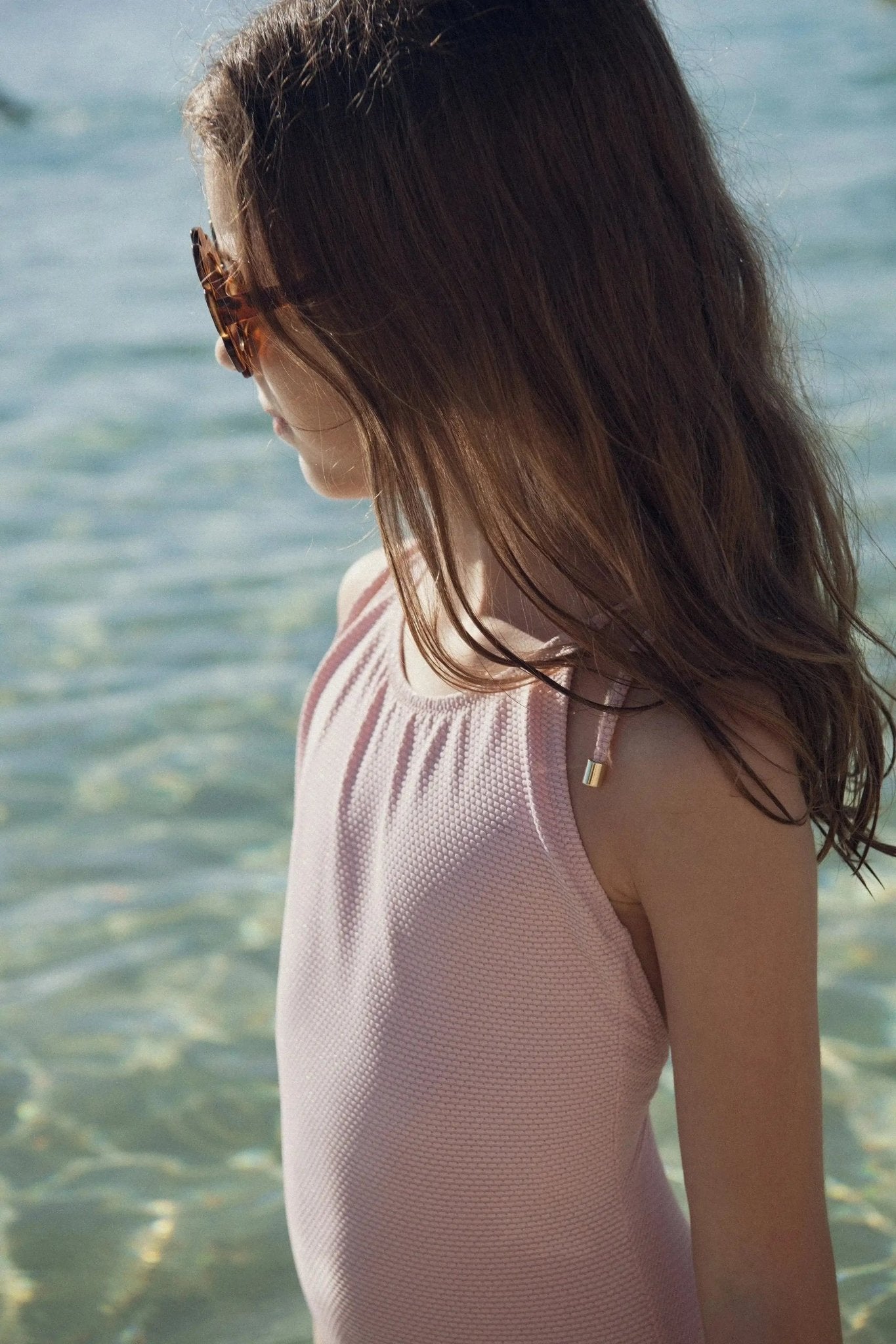 Girl in pale pink textured one-piece swimsuit standing by clear water, wearing sunglasses