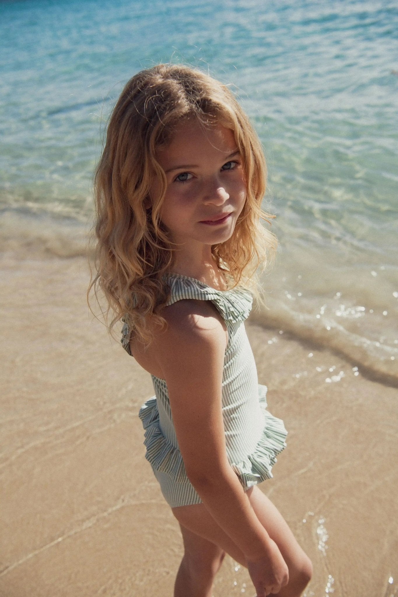 Girl in green striped one-piece swimsuit with ruffles standing by the sea on the beach