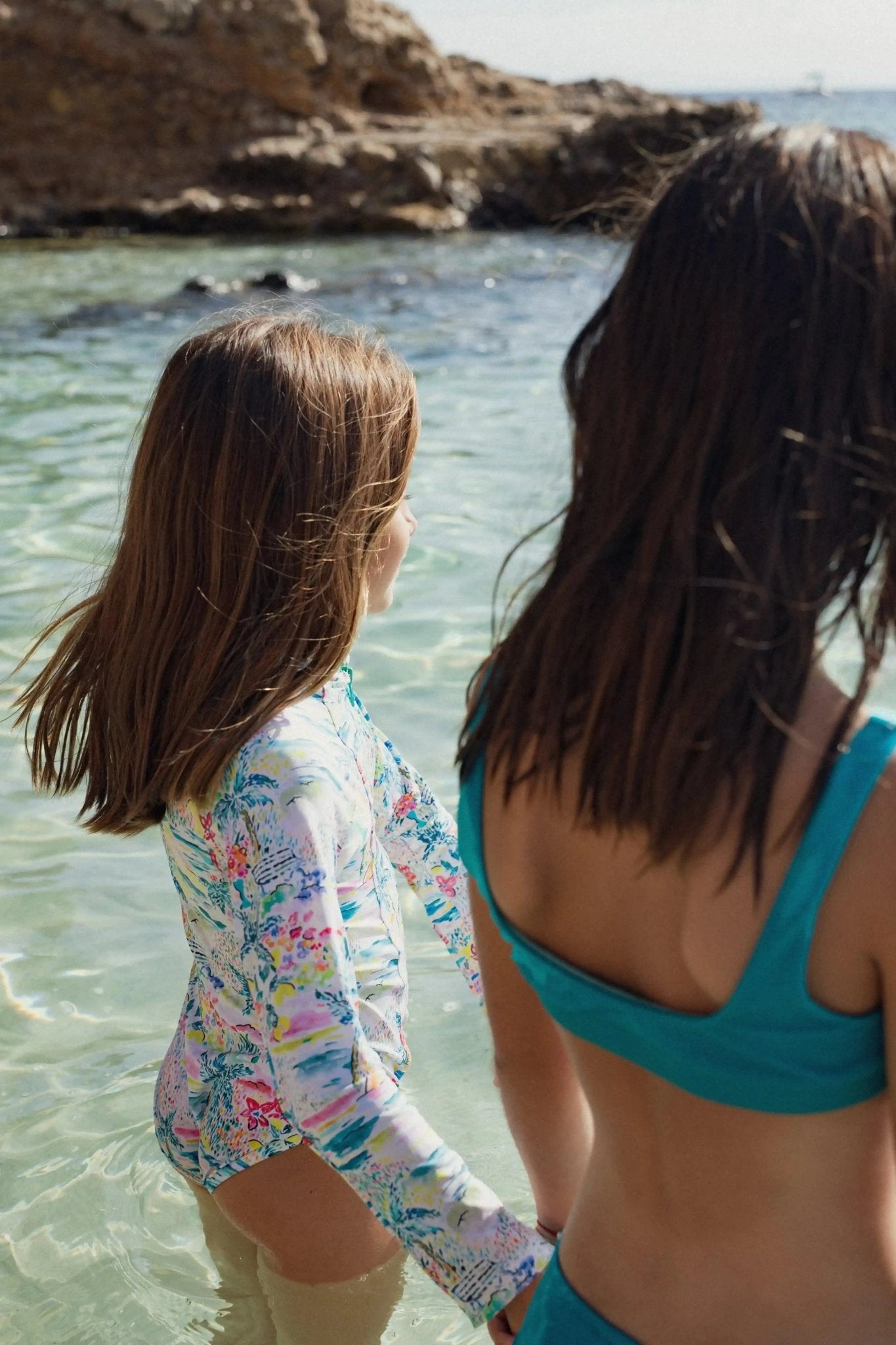 Two girls in colorful swimsuits wading in clear water at a rocky seaside beach