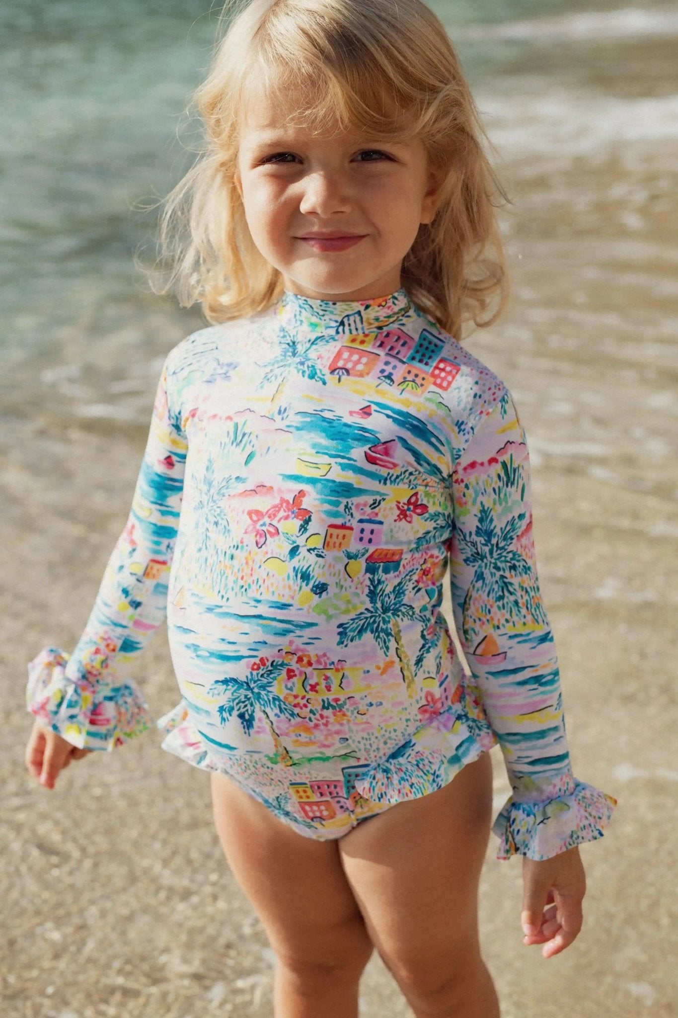 Young child at the beach wearing a colorful long-sleeve one-piece swimsuit with ruffles