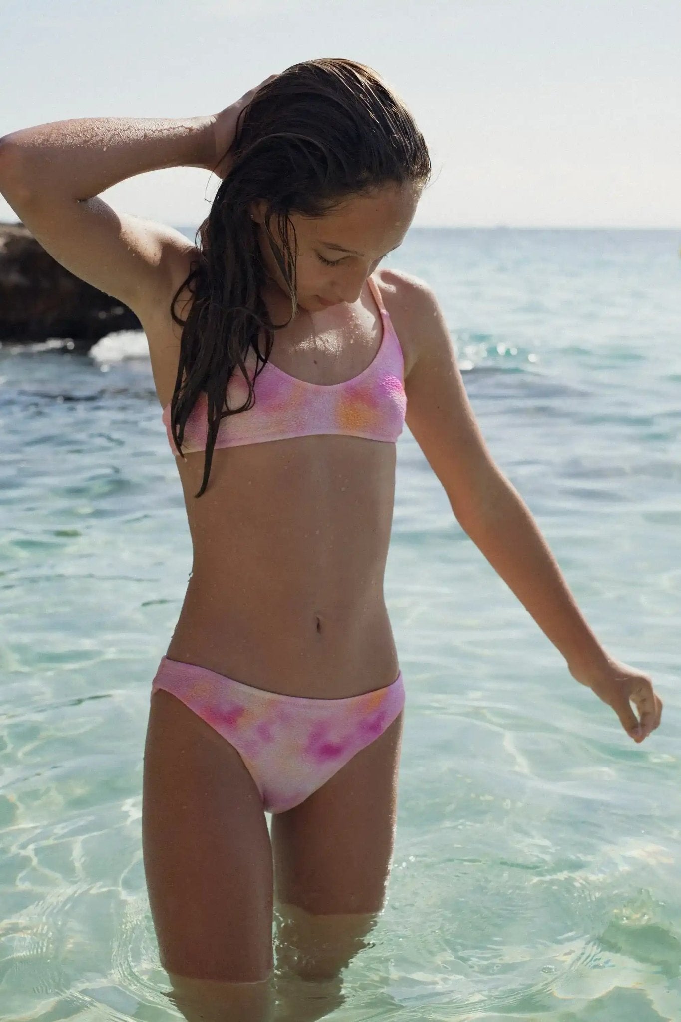 Girl wearing a pink and orange tie-dye two-piece swimsuit standing in clear ocean water