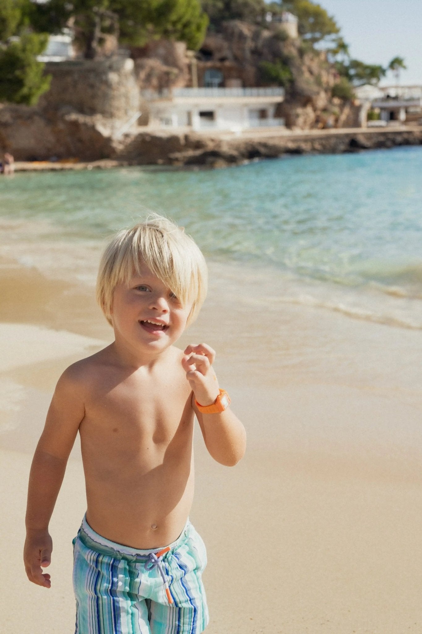 Young boy in striped swim shorts on a sunny beach near clear blue sea