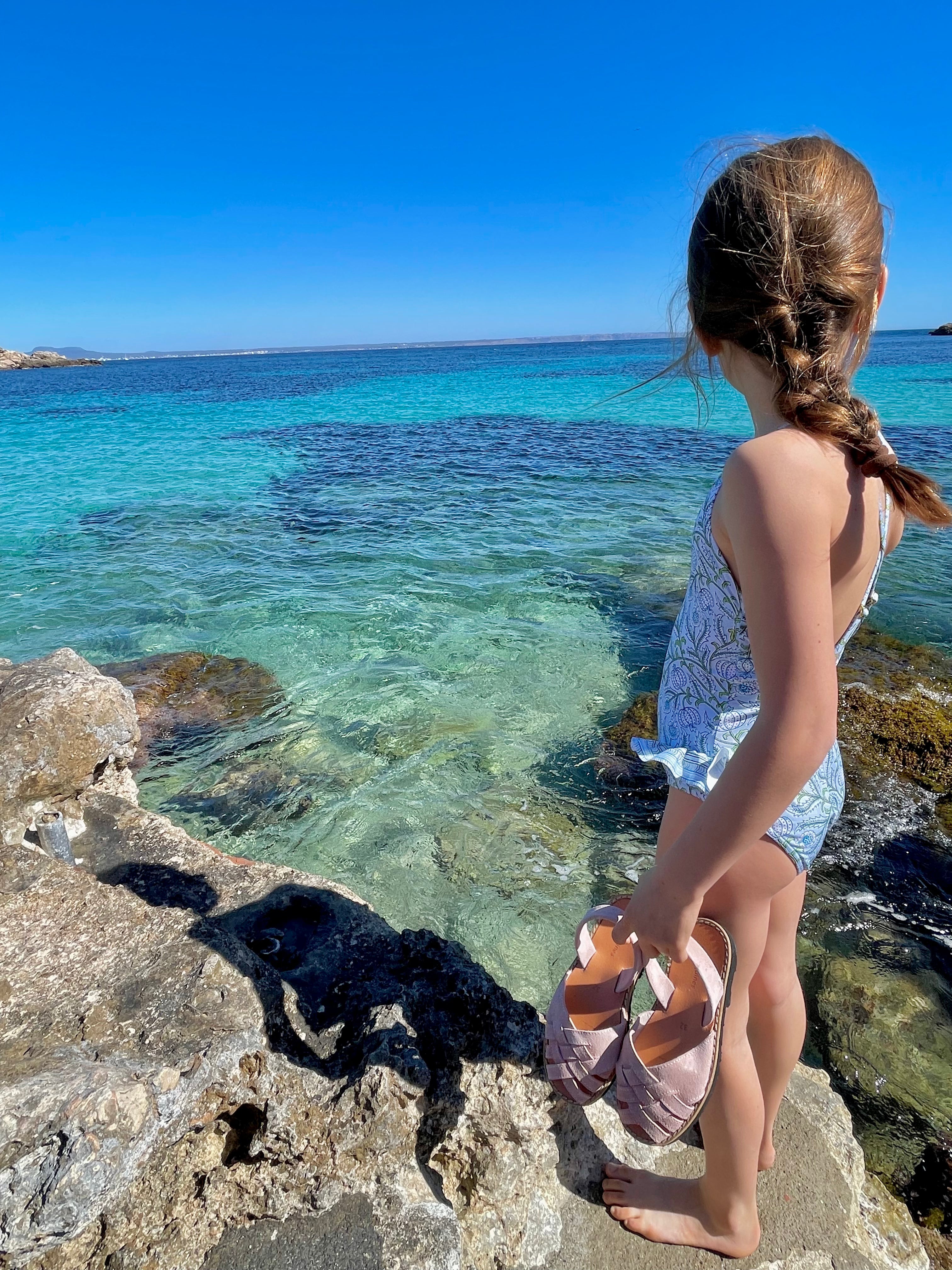 Girl in Lison Paris UV swimsuit holding pink sandals by the sea on a sunny beach day